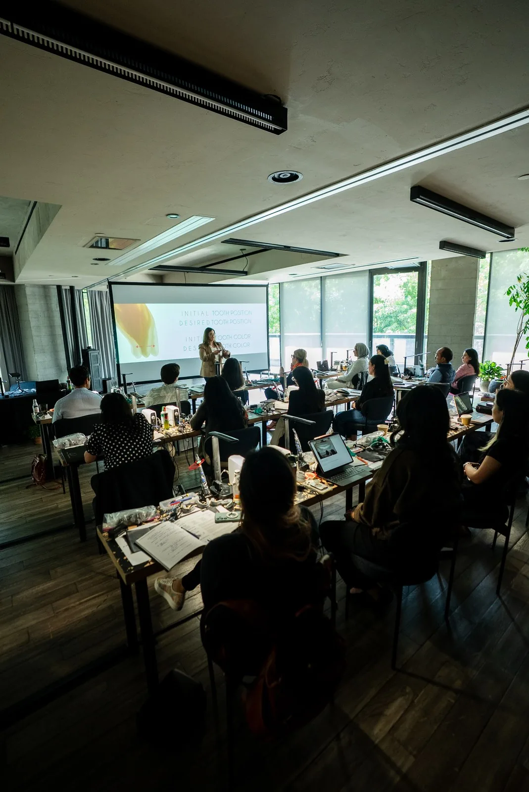 Dentists at instrument-covered benches watch a lecture slide on tooth position and color