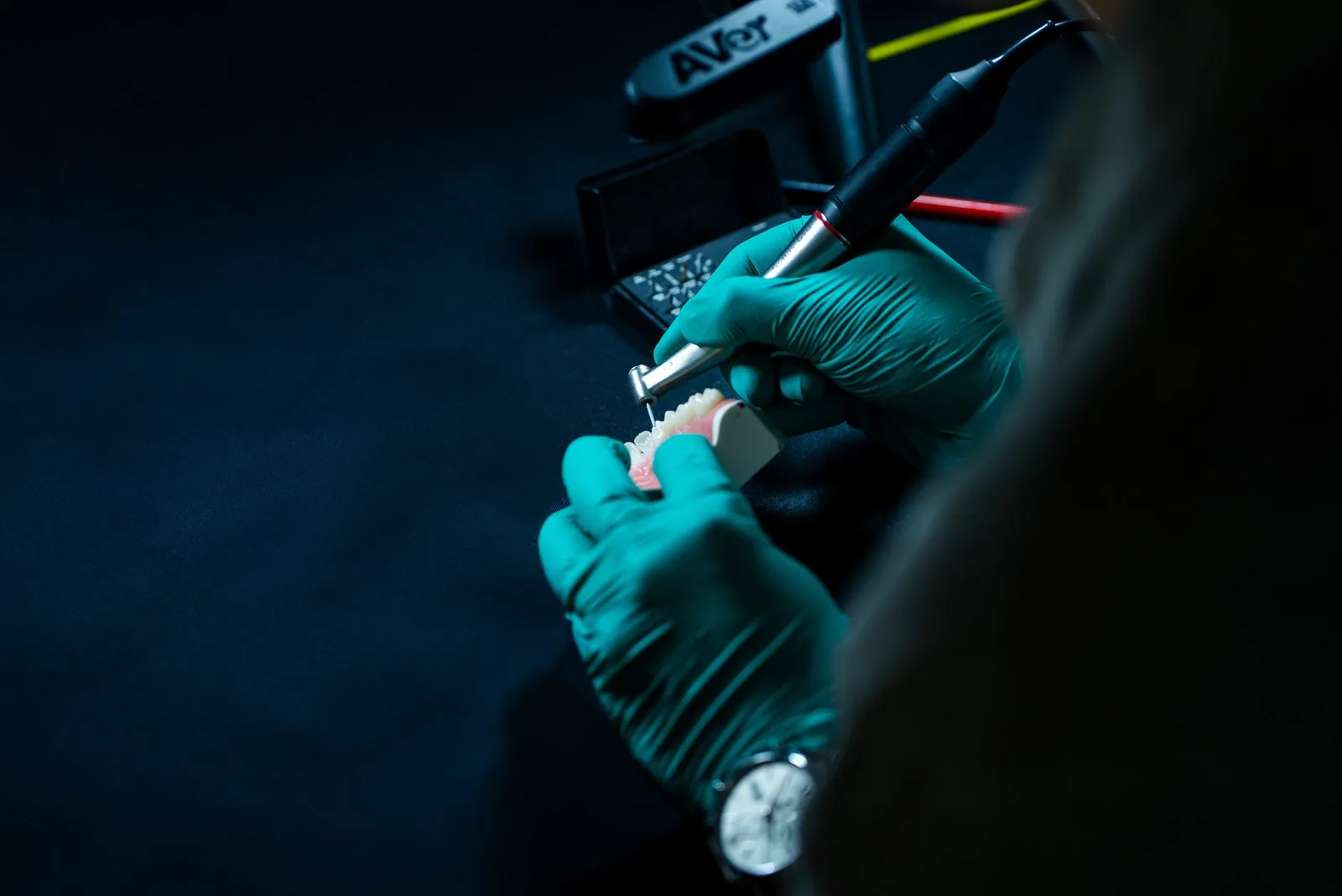 Teal-gloved hands shape a tooth on a typodont with a handpiece in low light